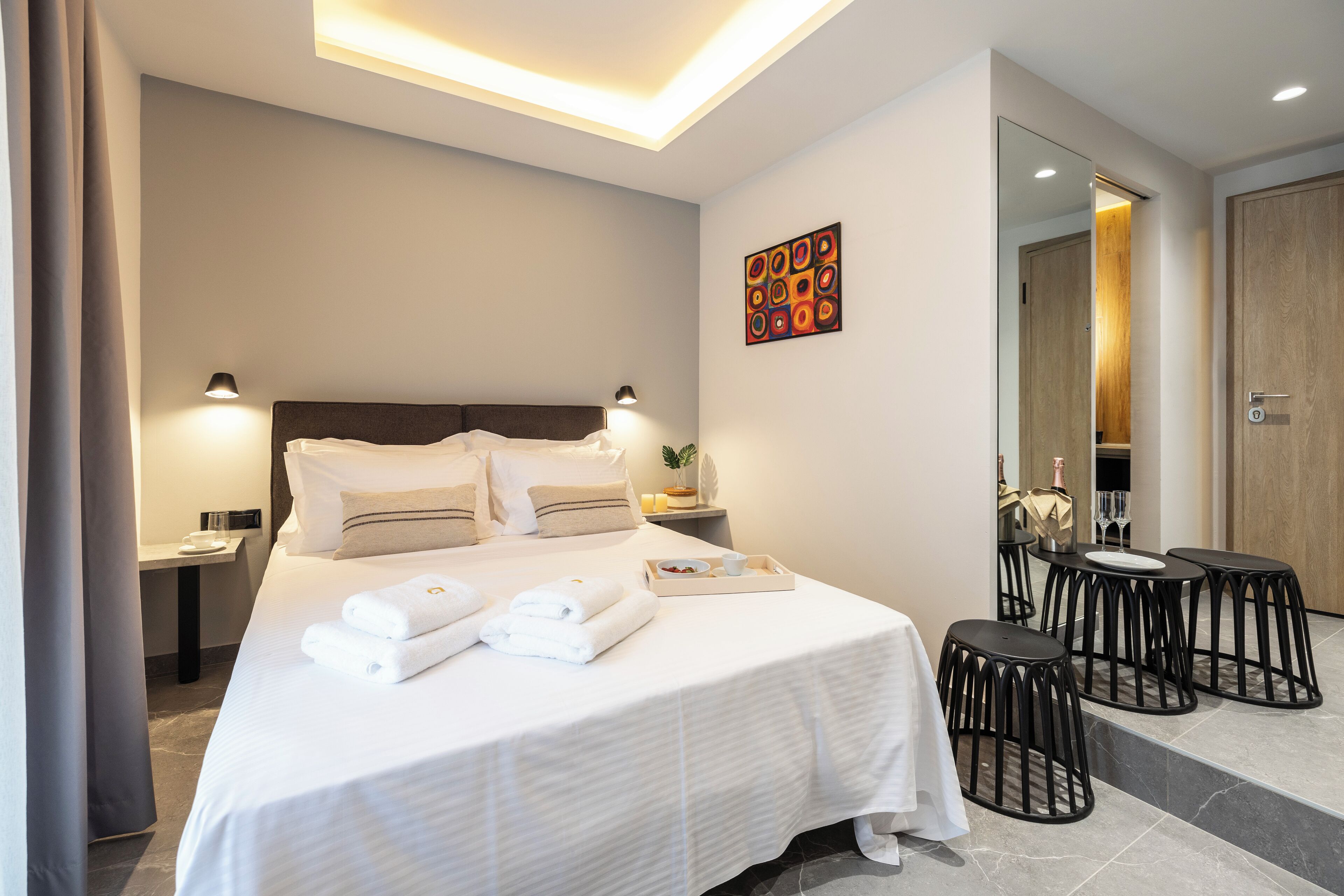 A hotel room with a neatly made bed, a small table with a tray of towels, and a seating area with stools.
