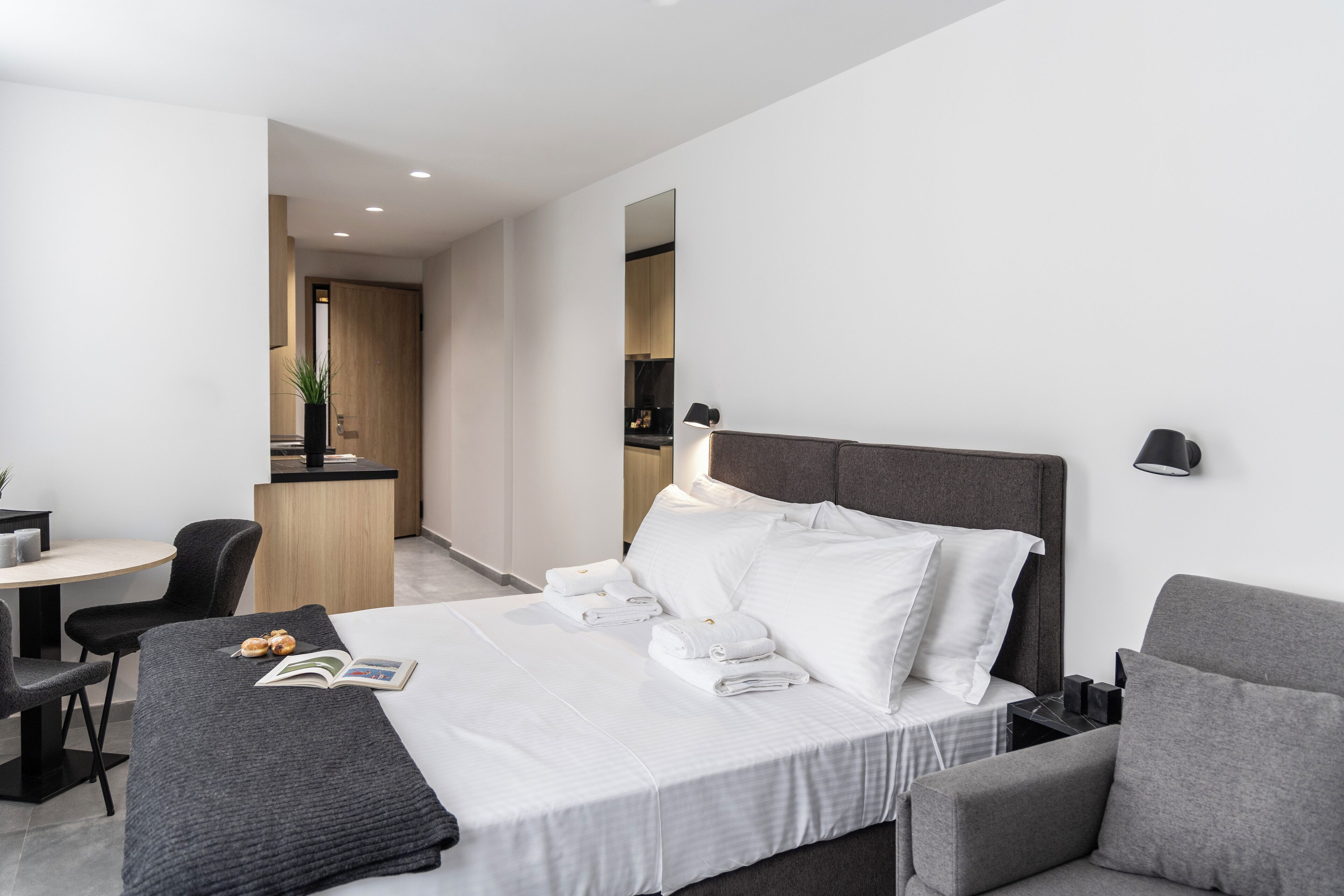 A modern hotel room with a neatly made bed, a grey sofa, a small table with a chair, and a view into the kitchen area.