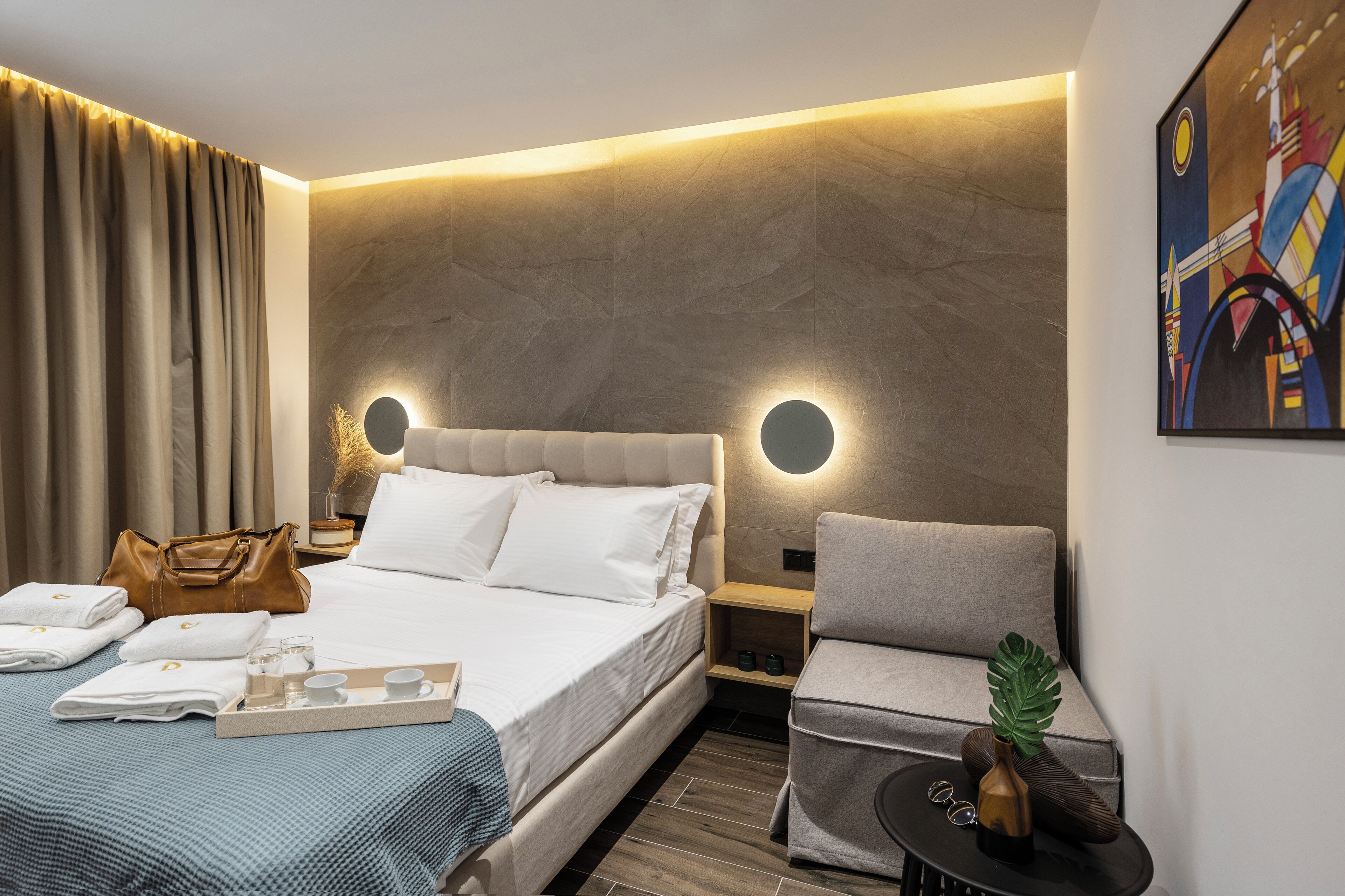 A modern hotel room with a bed, a chair, a bedside table, and a wall-mounted artwork.