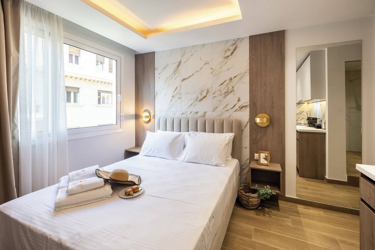 A modern hotel room with a large bed, a wooden headboard, a marble wall, and a view of the city.