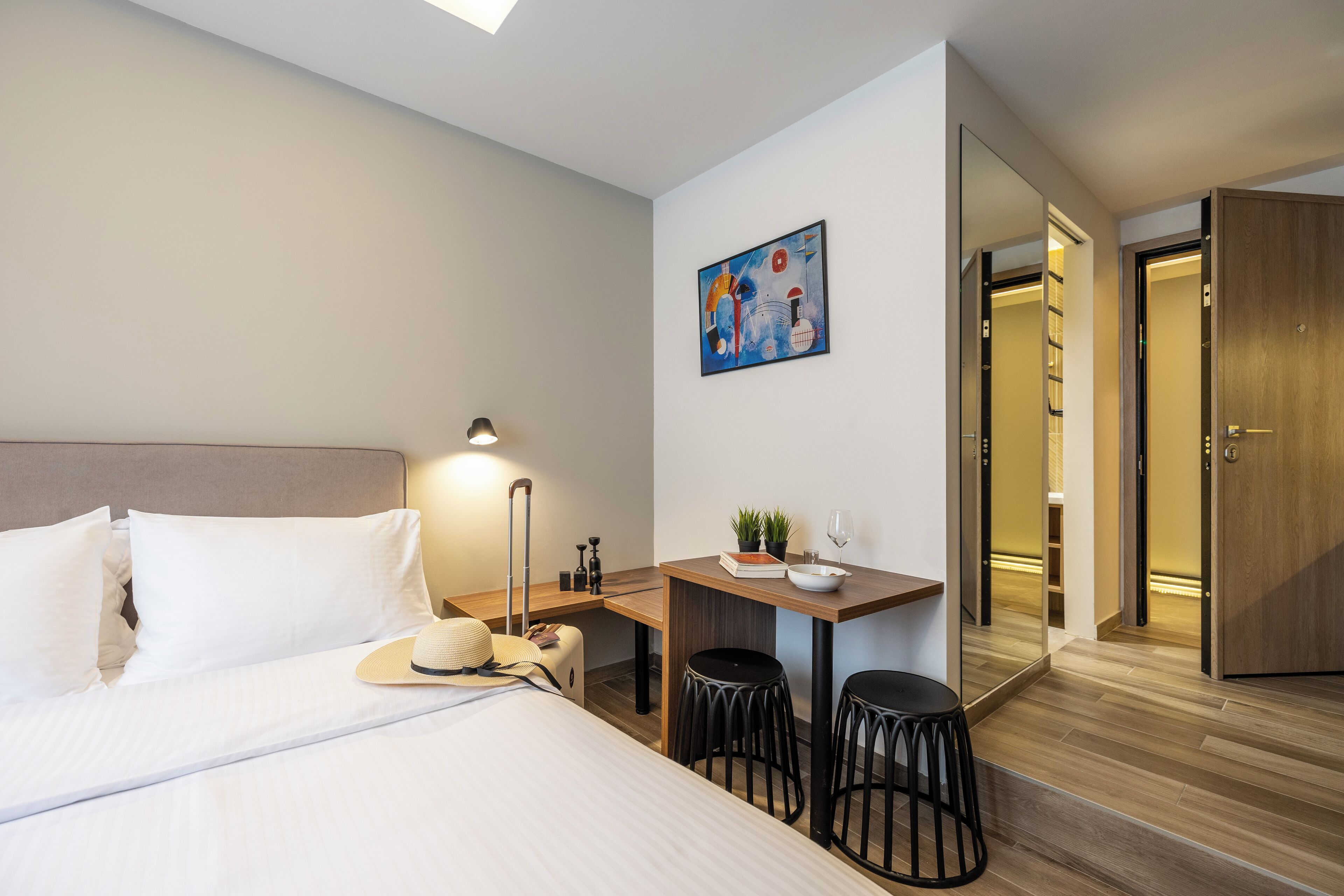 A hotel room with a bed, a desk, two stools, a small table with a plant and a wine glass, and a painting on the wall.