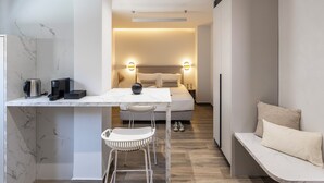 A modern hotel room with a marble-tiled wall, a white bed with a headboard, bedside lamps, a small table with a kettle and a coffee maker, and a bench with a pillow. - DOMIOS Suites Athens (Athens)