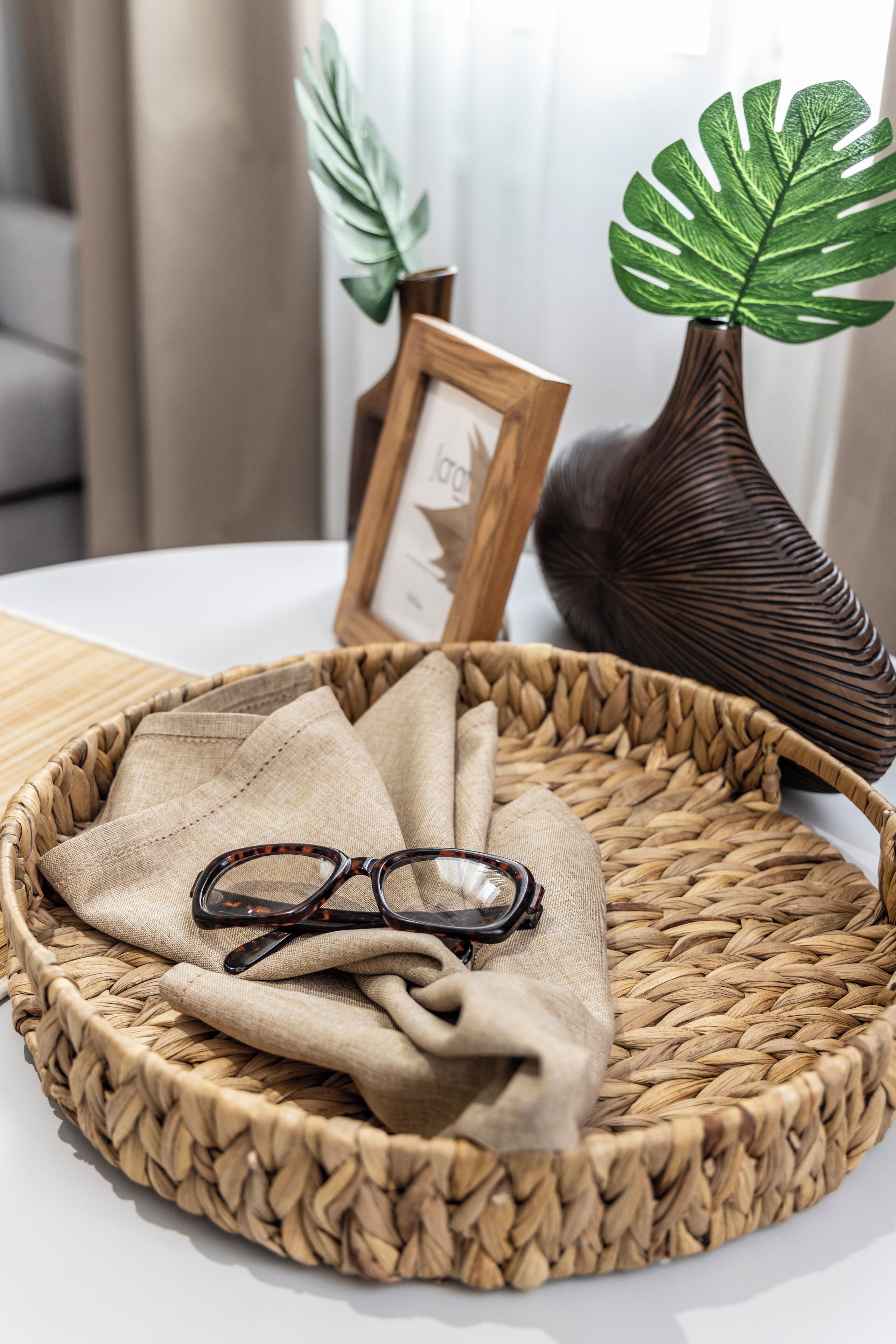 A woven basket with glasses and a fabric on a table, a framed picture, and two potted plants.