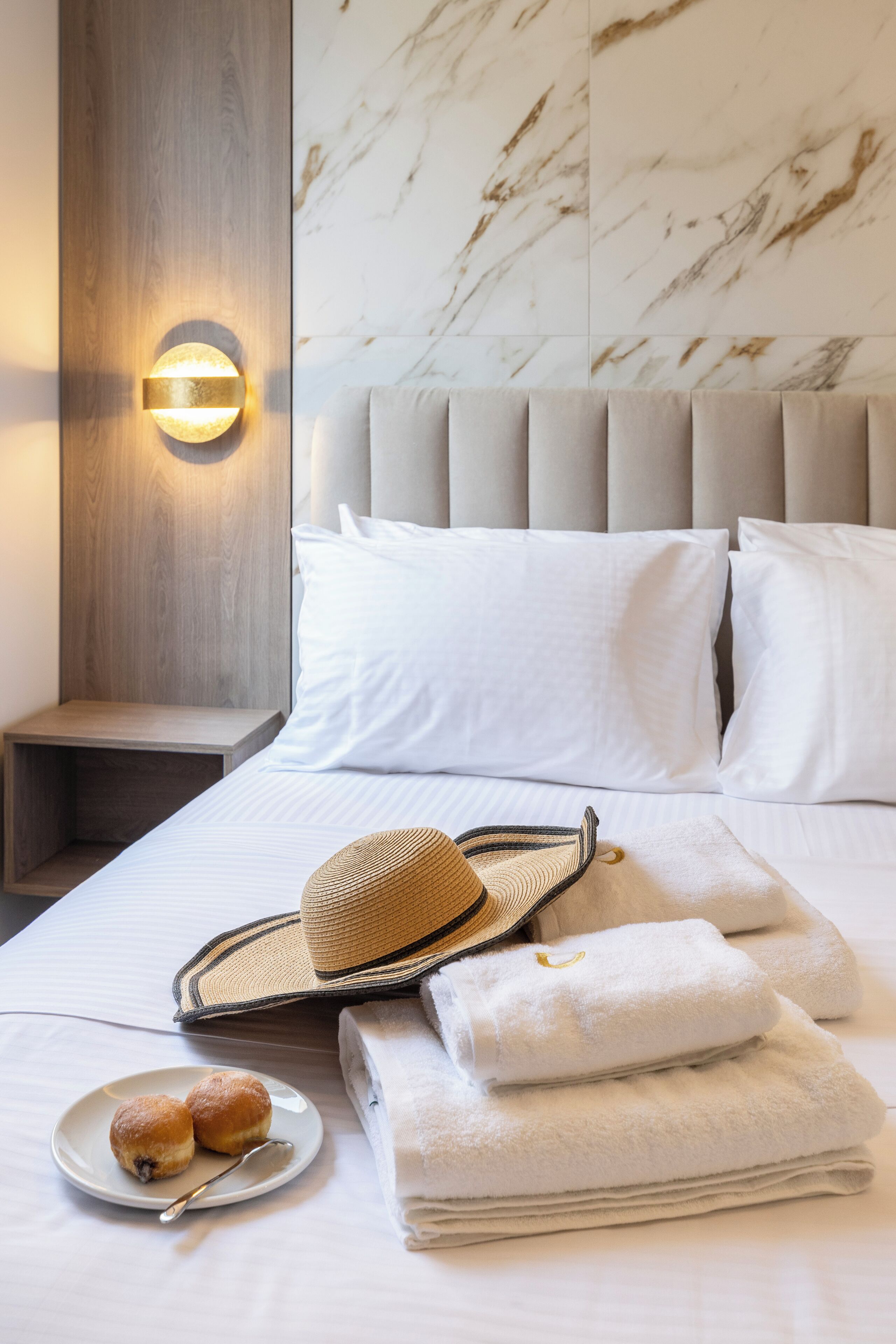 A neatly made bed with white linens, a bedside lamp, a woven hat, folded towels, and a plate with two pastries.