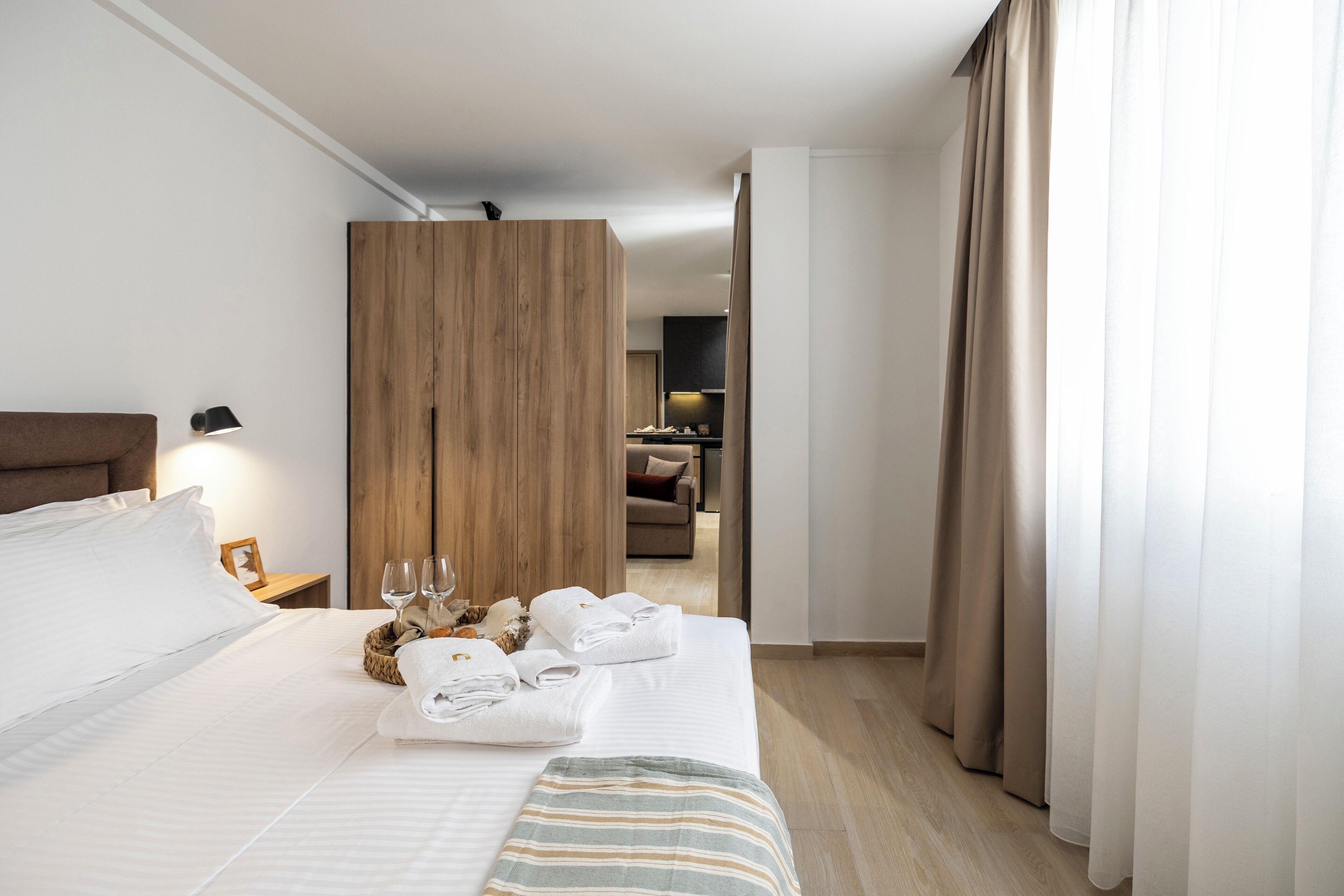A hotel room with a neatly made bed, a wooden wardrobe, a small table with a lamp, and a view of the hallway and another room.