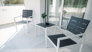 Een balkon met twee witte metalen stoelen en een kleine tafel, een vaas met bloemen, en een glazen deur die leidt naar een terras.