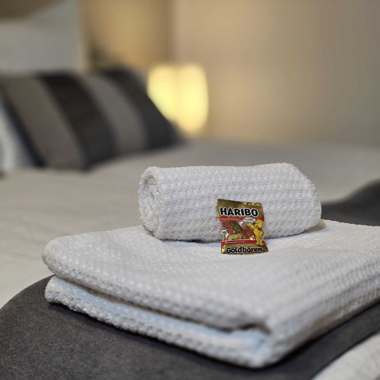 Una toalla blanca doblada con un paquete de Haribo Goldbären sobre una cama con ropa de cama gris y una almohada a rayas.