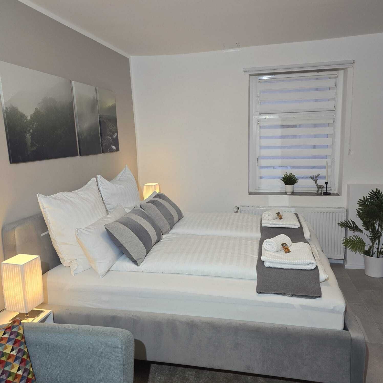 Une chambre bien rangée, avec un lit, des lampes de chevet, une fenêtre avec des stores, un radiateur et une plante en pot.
