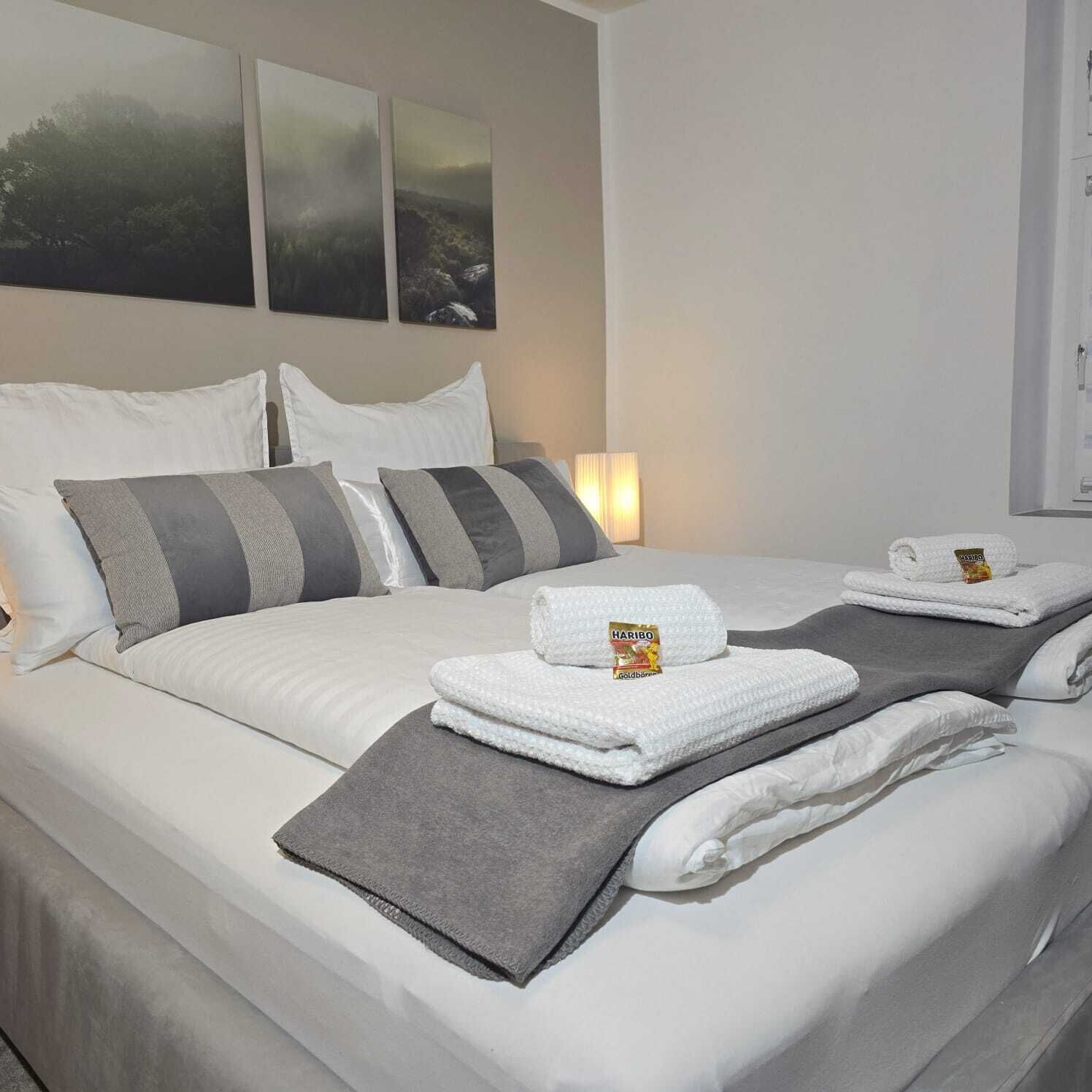 A hotel room with a neatly made bed, grey and white bedding, and a Haribo Goldbären package on the pillows.
