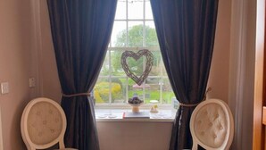 A room with two white upholstered chairs facing a window with dark curtains. A heart-shaped decoration hangs on the window, and a small table with a vase and a figurine is placed on the windowsill. - Charming Twin Room in a Unique and Beautiful B&B with Garden Views (Simonburn)