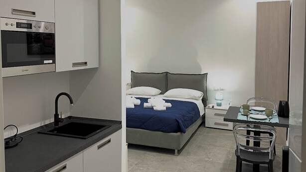 A modern hotel room with a kitchenette, a bed with blue bedding, a bedside table with a lamp, and a chair. - Cuore di Trastevere (Rome)
