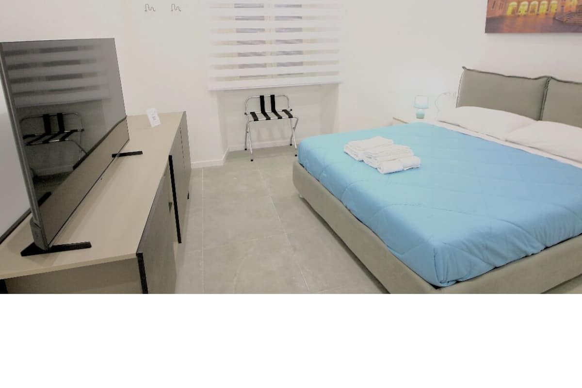 A bedroom with a bed, a desk, a chair, a window with blinds, and a painting on the wall.