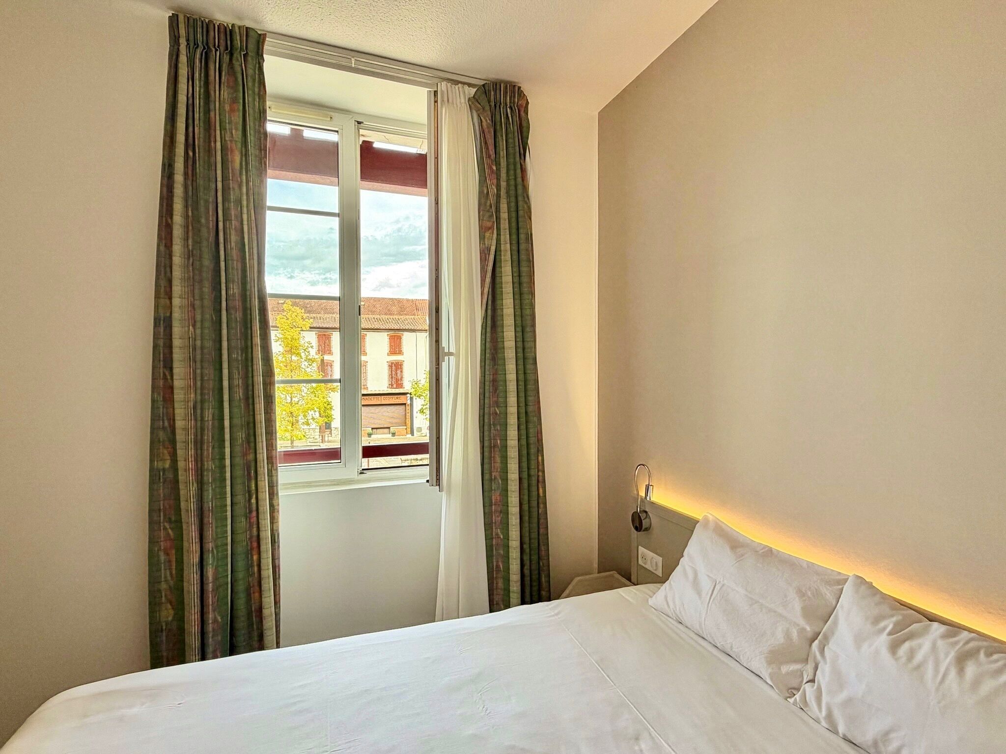 Una habitación de hotel con cama, cortinas, ventana y vistas a edificios.