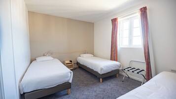A hotel room with two single beds, a small bedside table with a lamp, a window with curtains, and a radiator.
