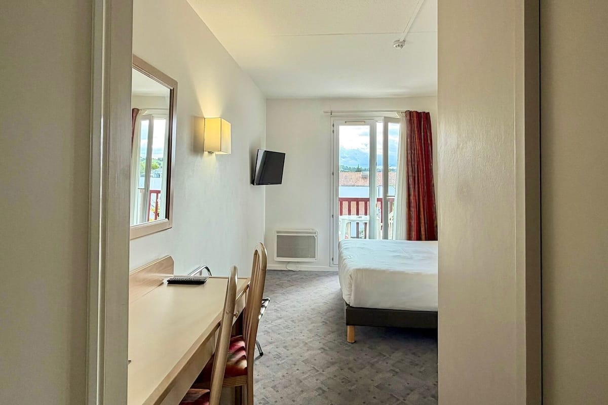 Une chambre d’hôtel comprenant un lit, un bureau avec une chaise, une télévision et une fenêtre avec des rideaux.