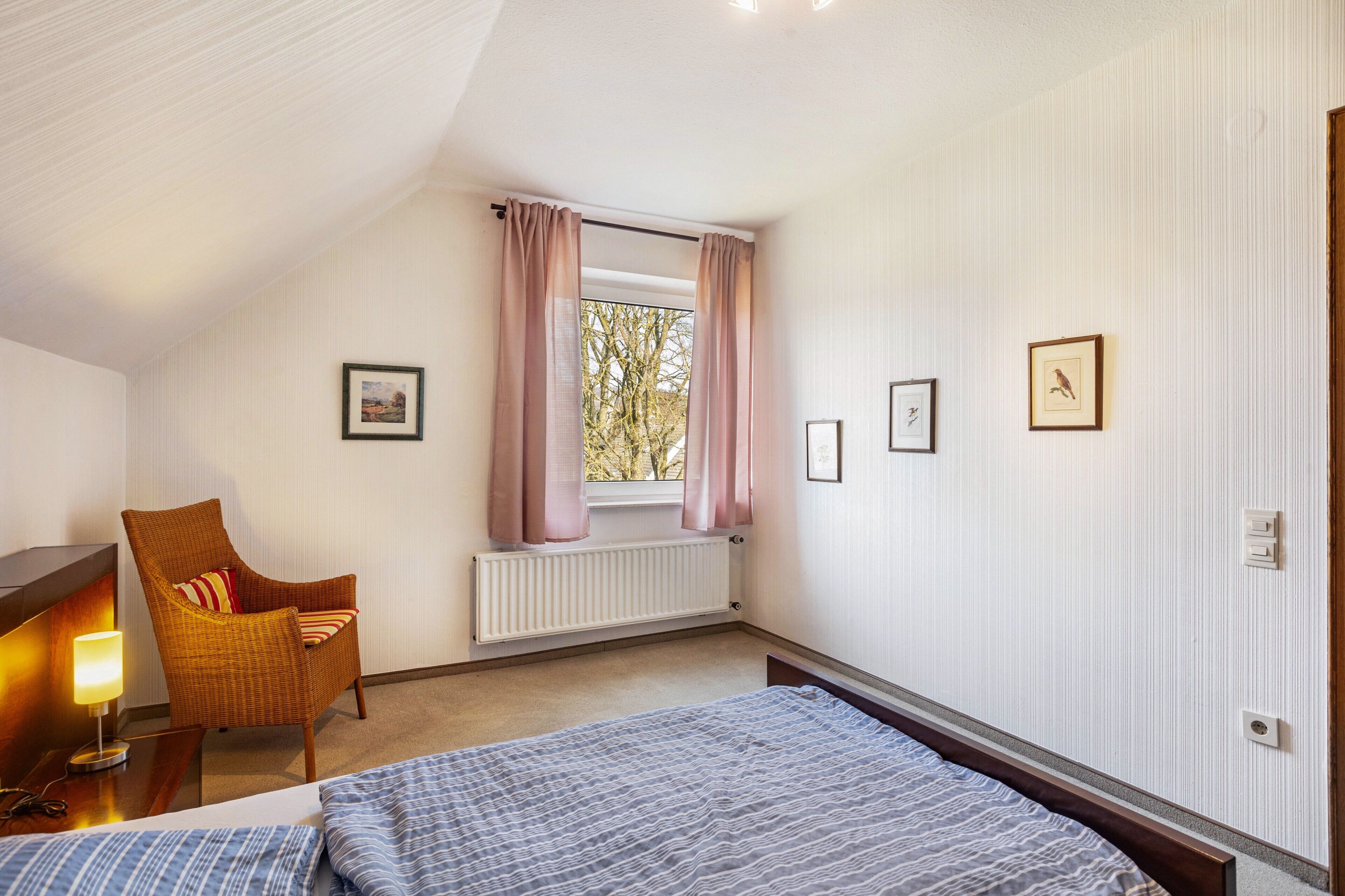 A bedroom with a bed, a chair, a window with curtains, a radiator, and framed pictures on the wall.