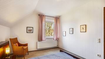 A bedroom with a bed, a chair, a window with curtains, a radiator, and framed pictures on the wall.