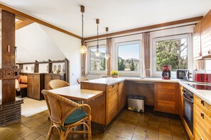 Eine Küche mit Holzausstattung, ein Essbereich mit Korbstühlen, ein Fenster mit Vorhängen und Blick auf Bäume.