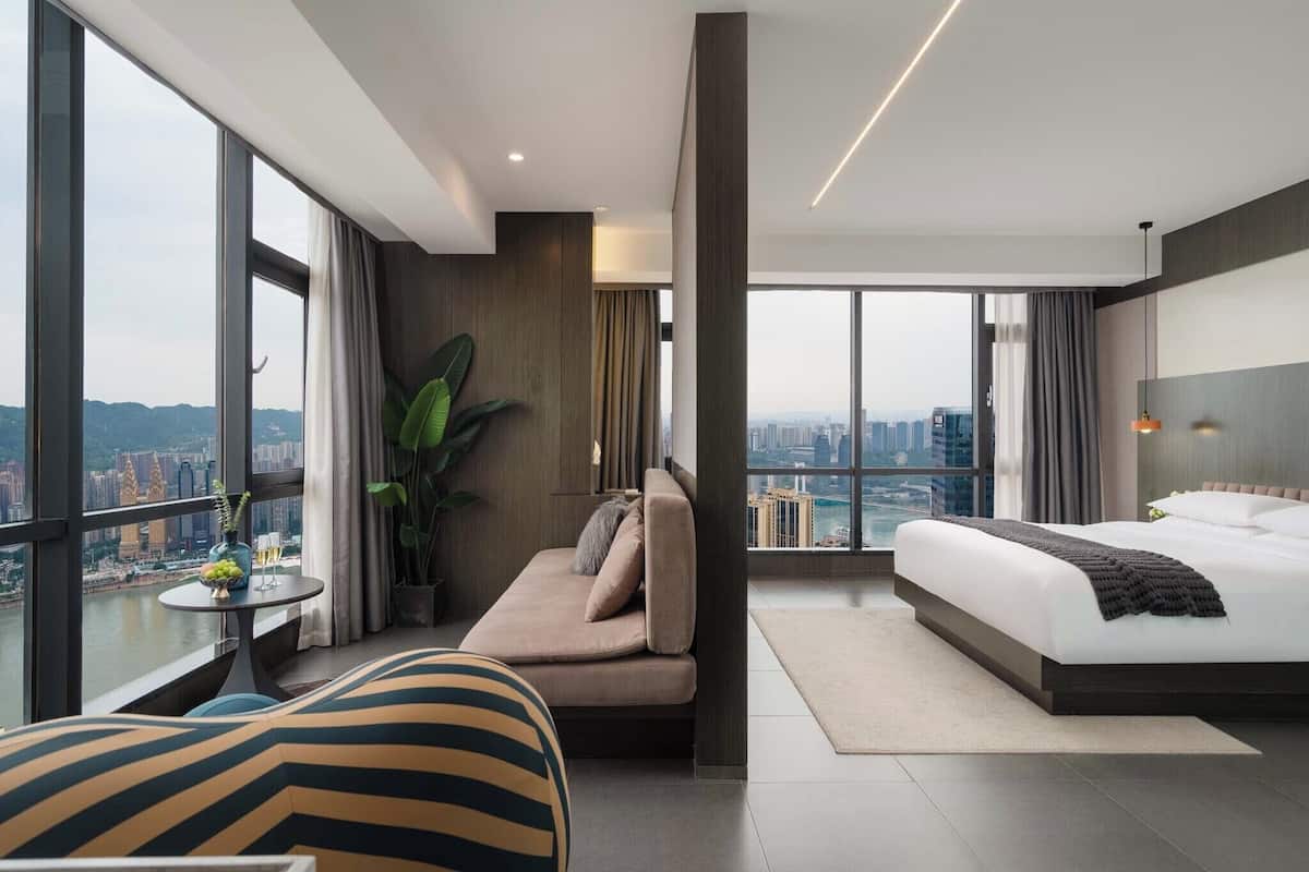 Habitación de hotel moderna con una cama grande, un sofá, una mesita con una planta y vistas a un paisaje urbano a través de amplios ventanales.
