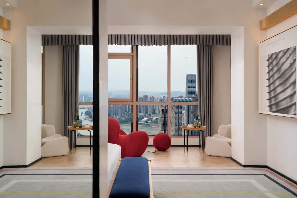 Una habitación de hotel moderna con un ventanal que ofrece vistas de la ciudad, un sofá, una silla roja, una mesita con un florero y un banco azul.
