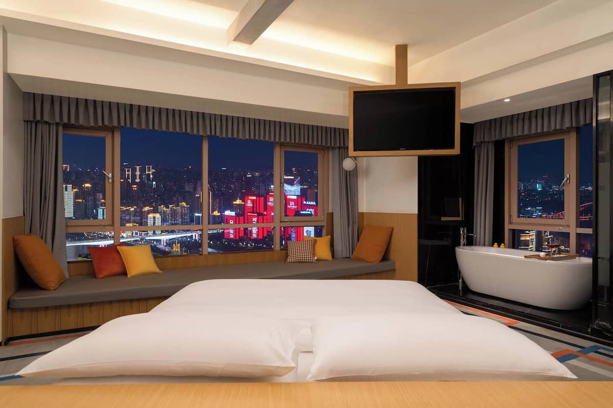 Habitación de hotel con una cama grande, una bañera y vistas a la ciudad a través de amplios ventanales.