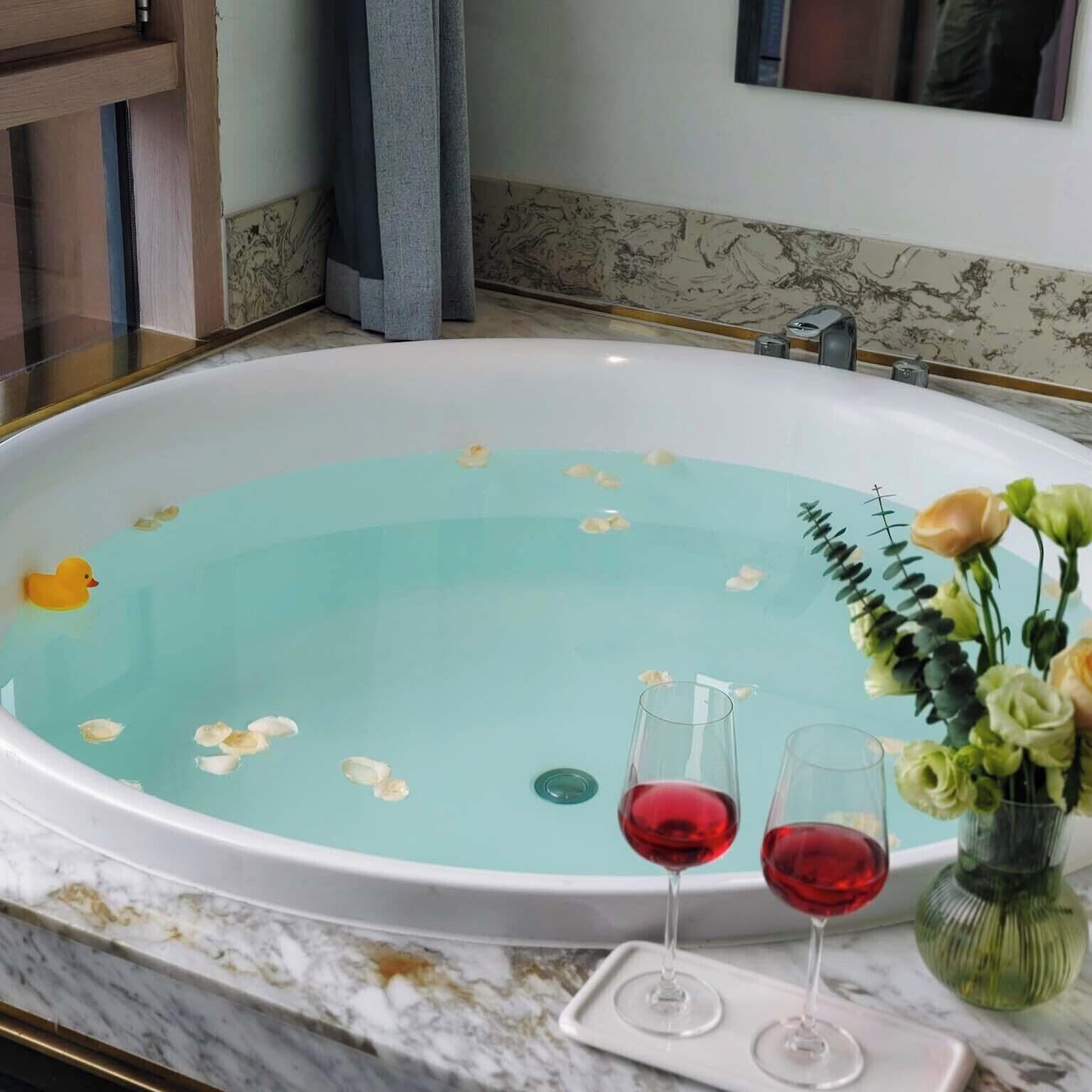A large bathtub with water, floating flowers, and a duck toy. There are two glasses of red wine on a tray next to the bathtub.