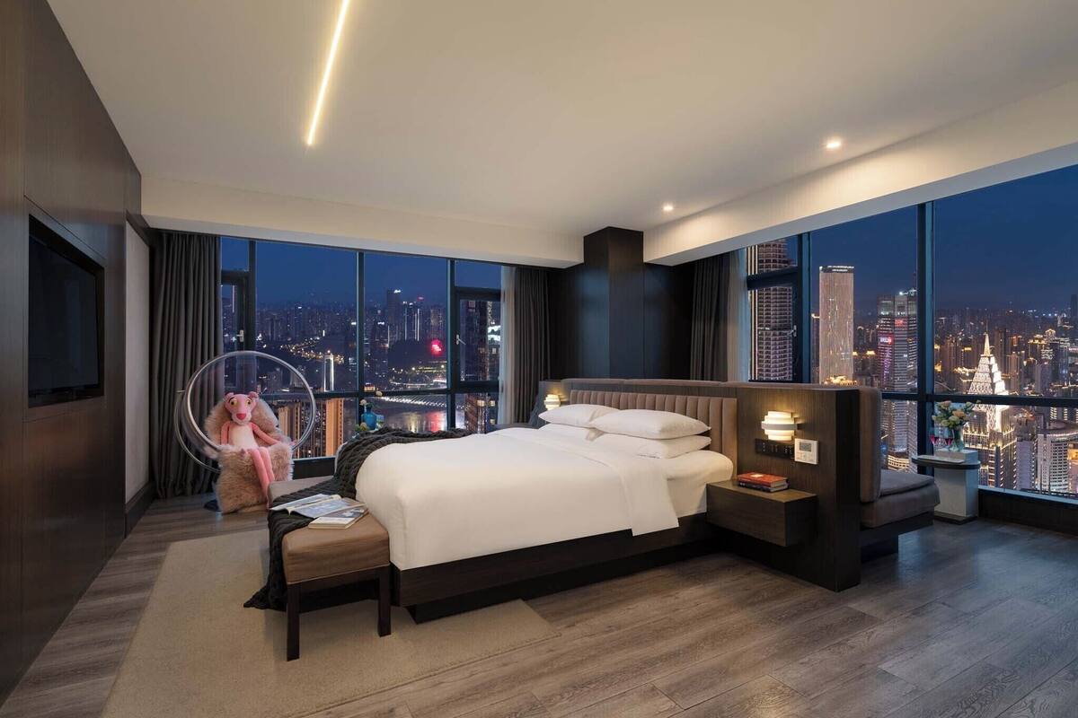 Habitación de hotel moderna con una cama grande, una mesita de noche y vistas a la ciudad iluminada de noche.