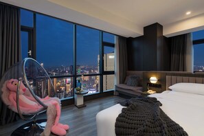 Una habitación de hotel moderna con una amplia vista de la ciudad, una cama acogedora, un sillón cómodo y una mesita de noche con lámpara.