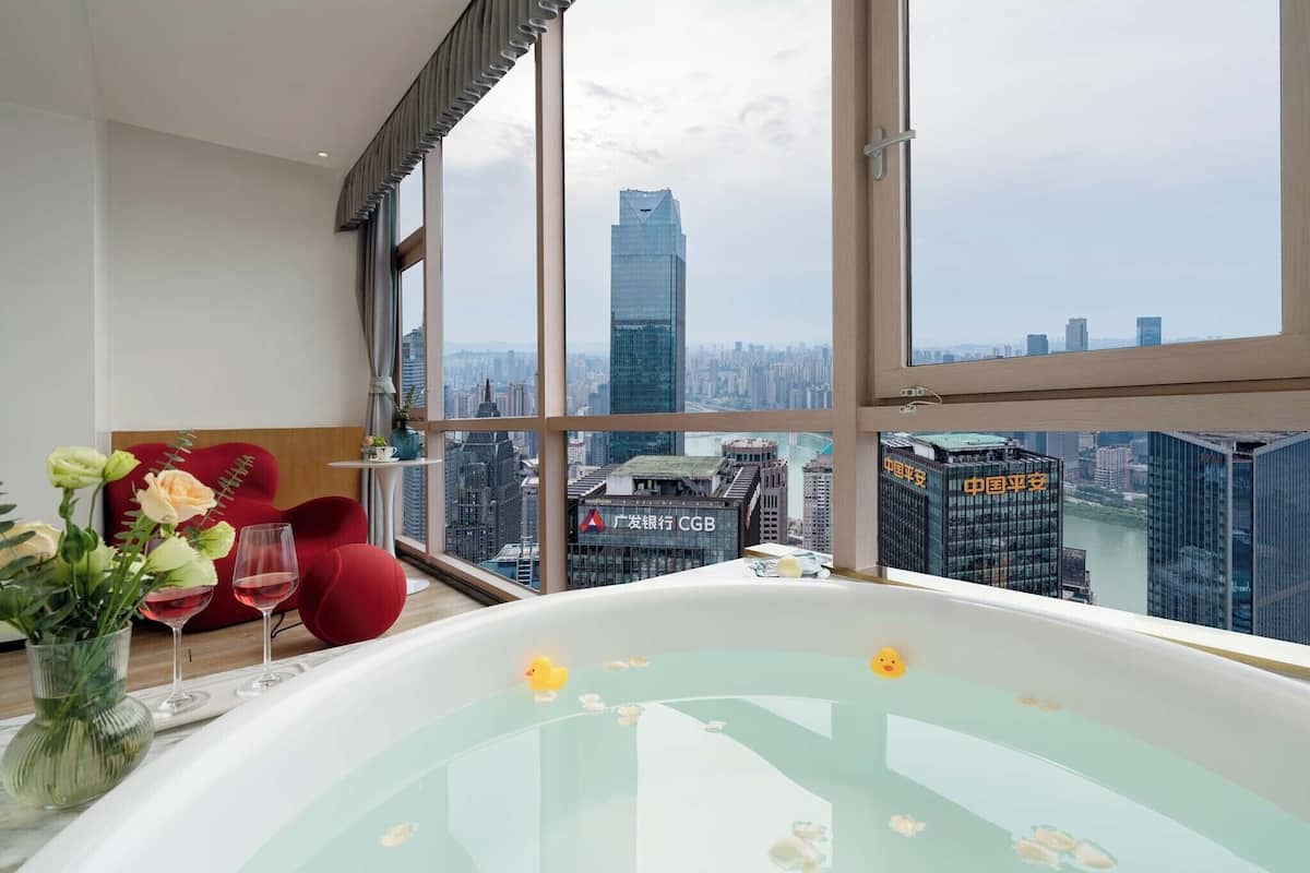 Una habitación moderna con un ventanal grande, una bañera con detalles florales, un florero con flores y dos copas de vino.