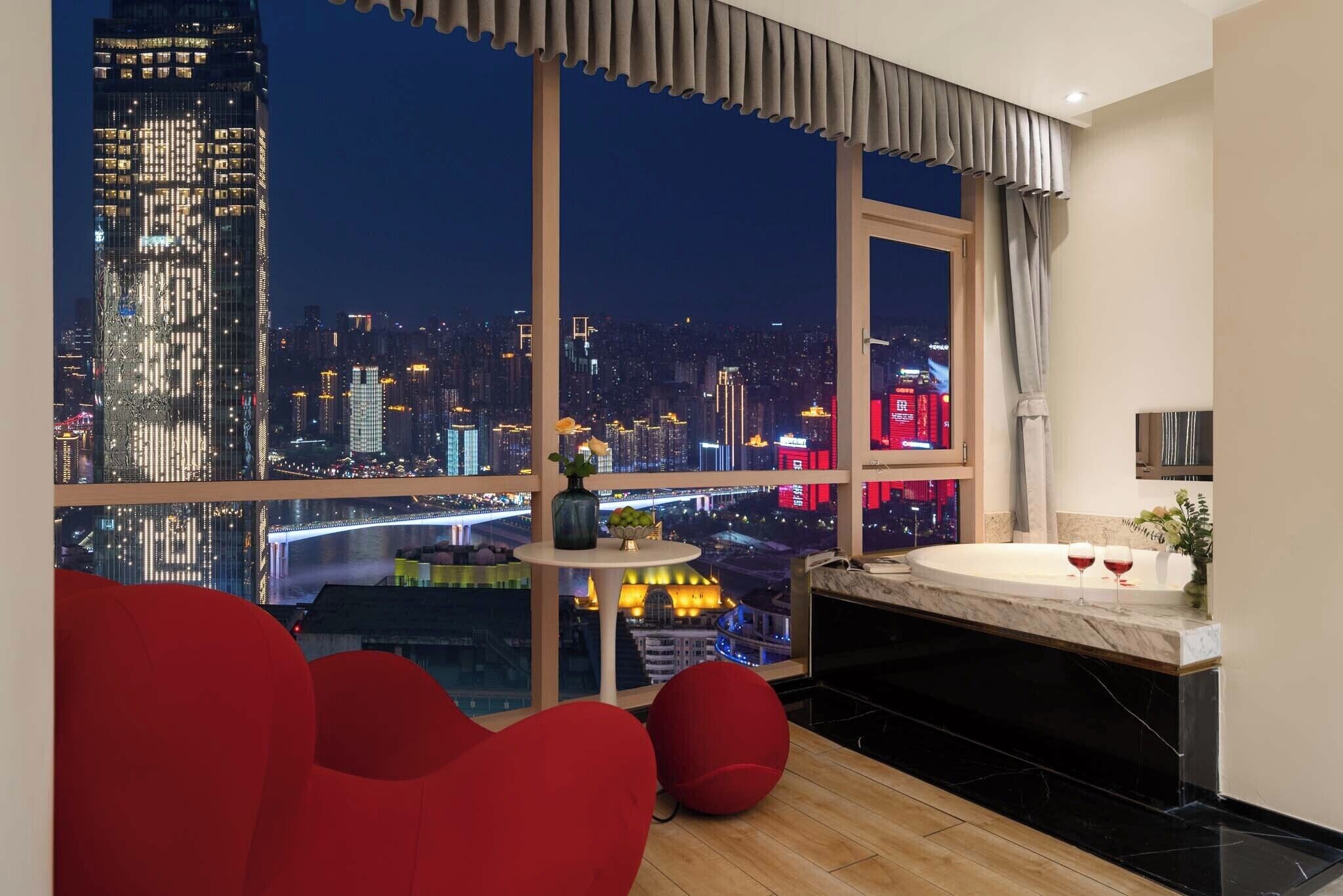 A modern hotel room with a large window offering a city view at night, a cozy seating area, and a marble bathtub.