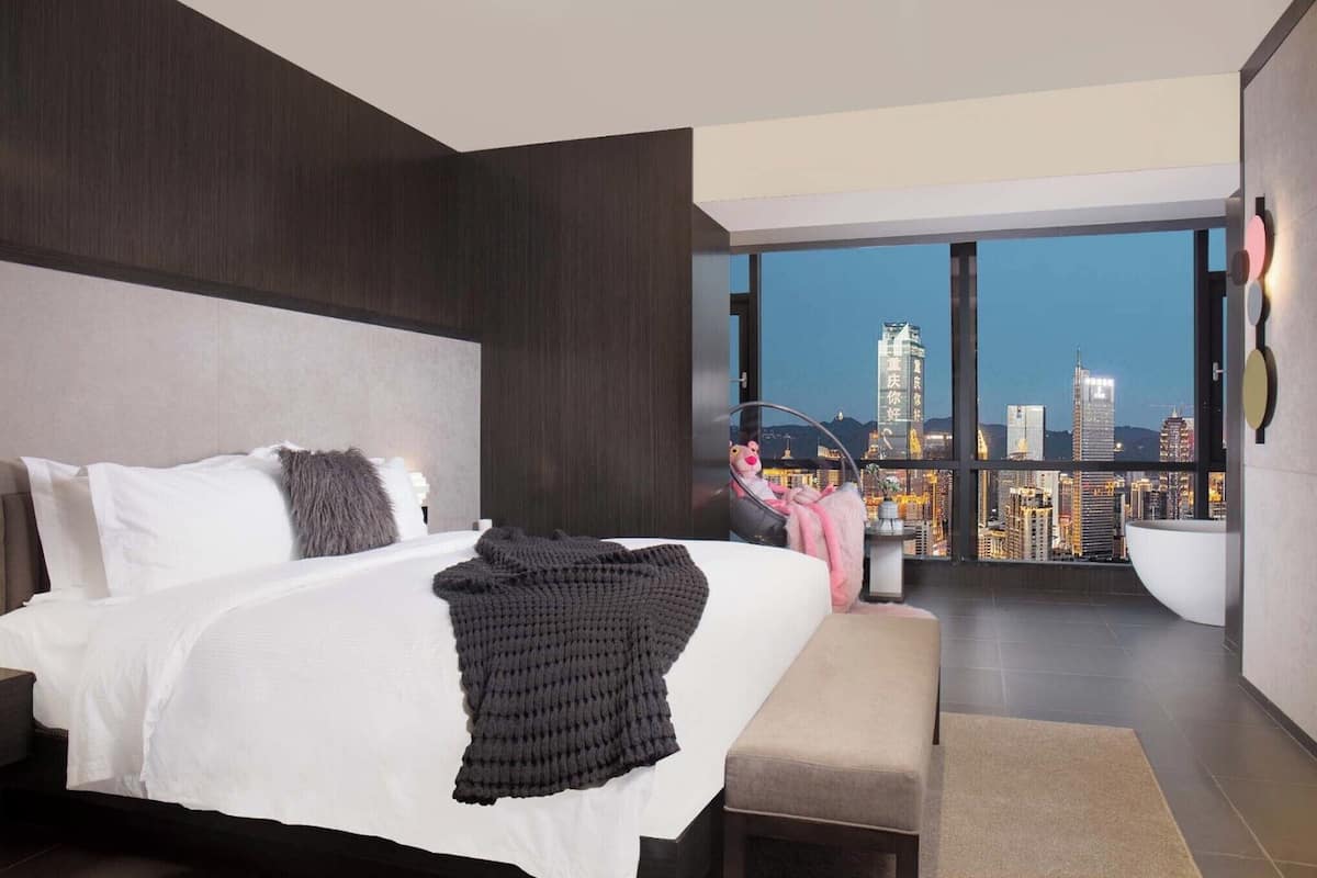 Habitación de hotel moderna con una cama grande, vistas a la ciudad y un balcón con zona de estar.