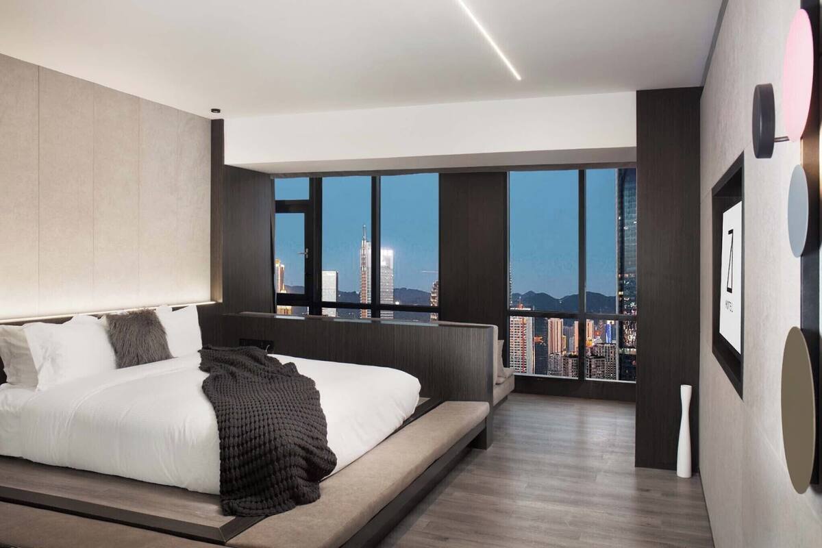 Un dormitorio moderno con una cama grande, vistas de la ciudad a través de ventanas de piso a techo y una decoración minimalista.