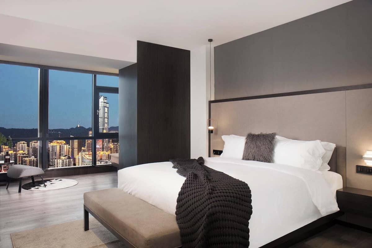 Habitación de hotel moderna con una cama grande, un banco y vista a la ciudad a través de amplios ventanales.