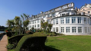 Un grand bâtiment blanc à plusieurs étages, doté de balcons et de grandes fenêtres, entouré de jardins bien entretenus et d’un sentier.