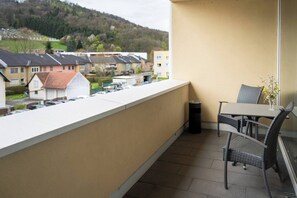 Un balcon avec une table et des chaises, un vase rempli de fleurs et une vue sur des maisons et une colline en arrière-plan.