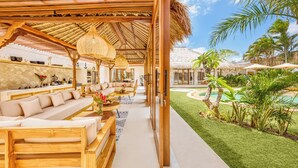 Amplia zona de estar al aire libre con mobiliario de madera, techo de paja y vista a una piscina y jardín tropical.
