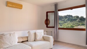 A modern living room with a white sofa, a large window with a view of greenery, a radiator, and a wall-mounted air conditioner. - Casa Leo by Wonderful Italy (Olbia)