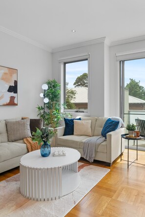 Ein modernes Wohnzimmer mit einem runden weißen Couchtisch, einem beige farbenen Sofa und einem großen Fenster mit Blick ins Freie.
