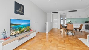 Un apartamento moderno con suelo de madera, zona de comedor con mesa y sillas, cocina con armarios blancos y un televisor mostrando un paisaje escénico.