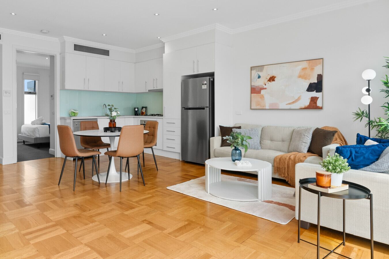 Una sala de estar moderna con un sofá, una mesa de centro y sillas. Se observa un comedor con una mesa y sillas. En el fondo, se ve una cocina con armarios y electrodomésticos blancos.