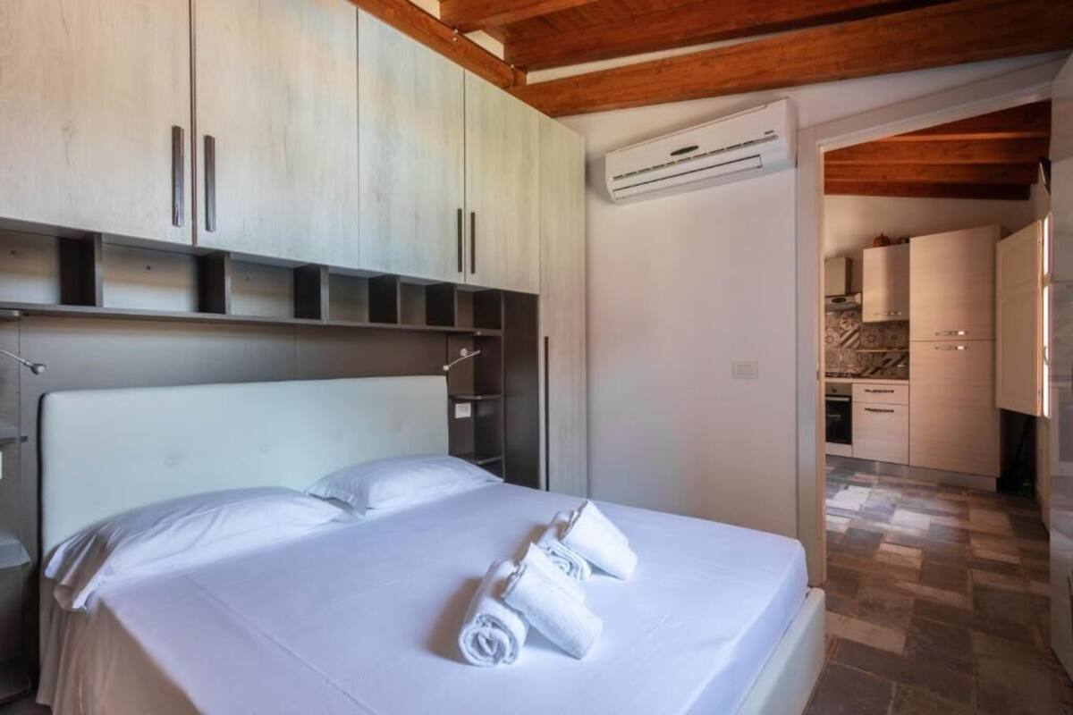 A bedroom with a bed, white linens, and a set of towels. There is a kitchenette visible in the background.