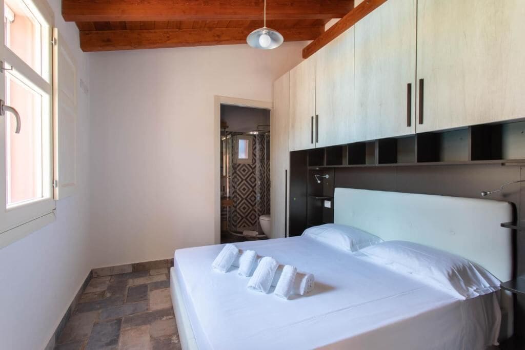 A bedroom with a bed, wooden ceiling beams, a wardrobe, and a bathroom visible through an open door.