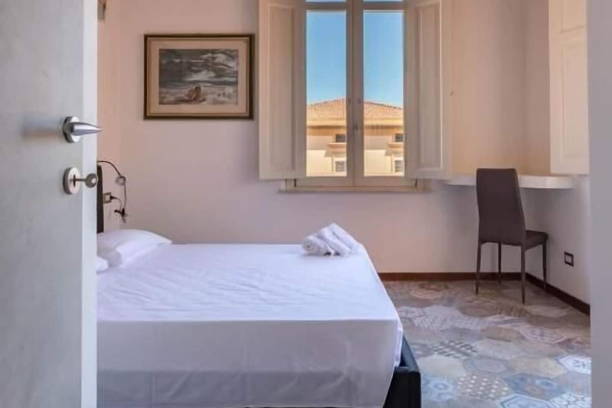 A bedroom with a wooden ceiling, a bed, a chair, a window with shutters, and a patterned floor.