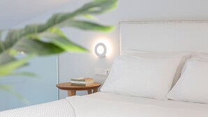 A neatly made bed with white linens, a round wall-mounted light, and a small wooden nightstand with a book. - Lirides Suites (Lefkada)