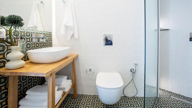 A bathroom with a large white sink, a toilet, and a shower area. There are towels on a shelf and a patterned floor. - Lirides Suites (Lefkada)