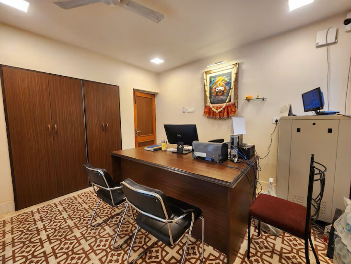 A reception desk with a computer, two chairs, a patterned carpet, a wooden wardrobe, and a framed picture on the wall.