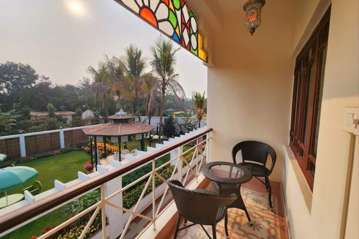 A balcony with a view of a garden, a small table, two chairs, and a decorative stained glass window.