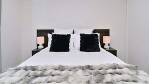 Una cama bien hecha con edredón blanco, dos almohadas negras y una manta suave gris y blanca. Hay dos mesitas de noche con lámparas a cada lado de la cama.
