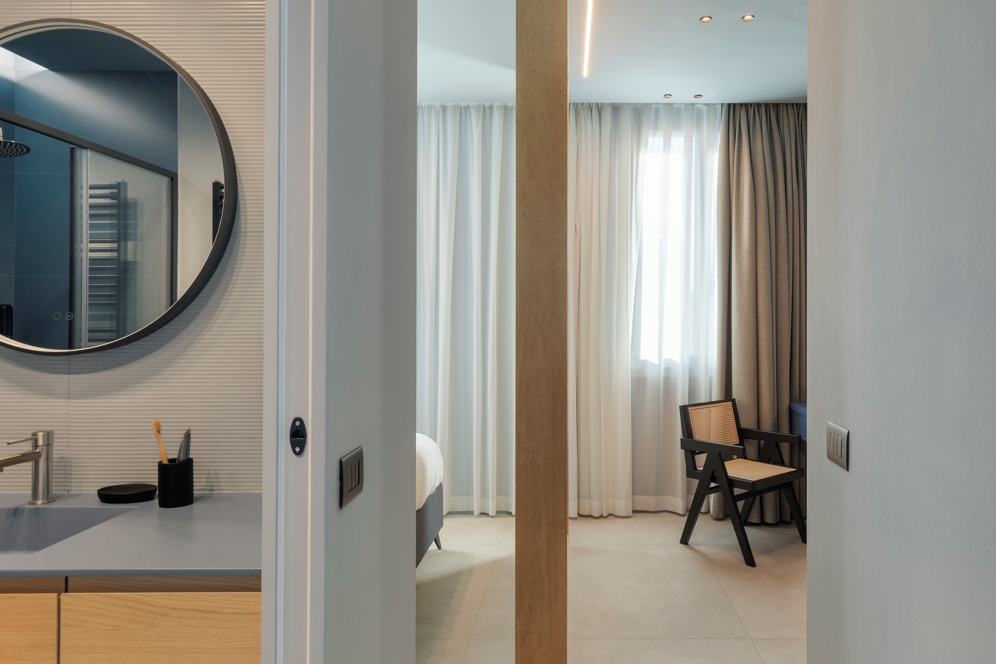 A modern hotel room with a round mirror, a bed, a chair, and a bathroom with a sink and towel rack.