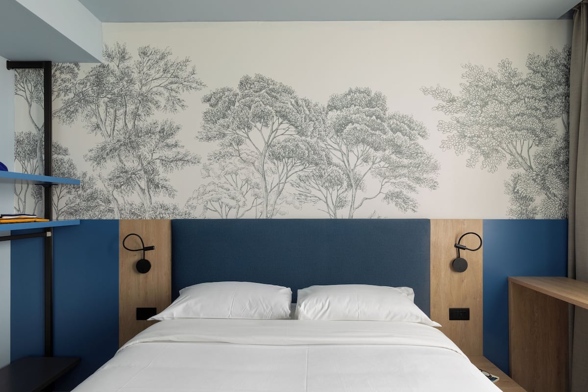 Une chambre avec un lit, une étagère fixée au mur et un mur décoré d’une fresque représentant un arbre.